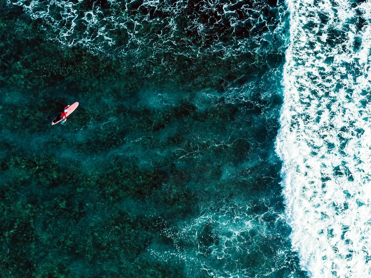Aerial view of a surfer paddling a red board over a turquoise reef beside breaking whitewater in Siargao
