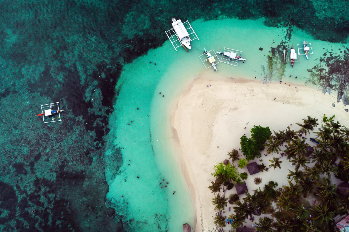 Aerial view of Guyam Island’s white sand and turquoise shallows, with outrigger boats moored nearby